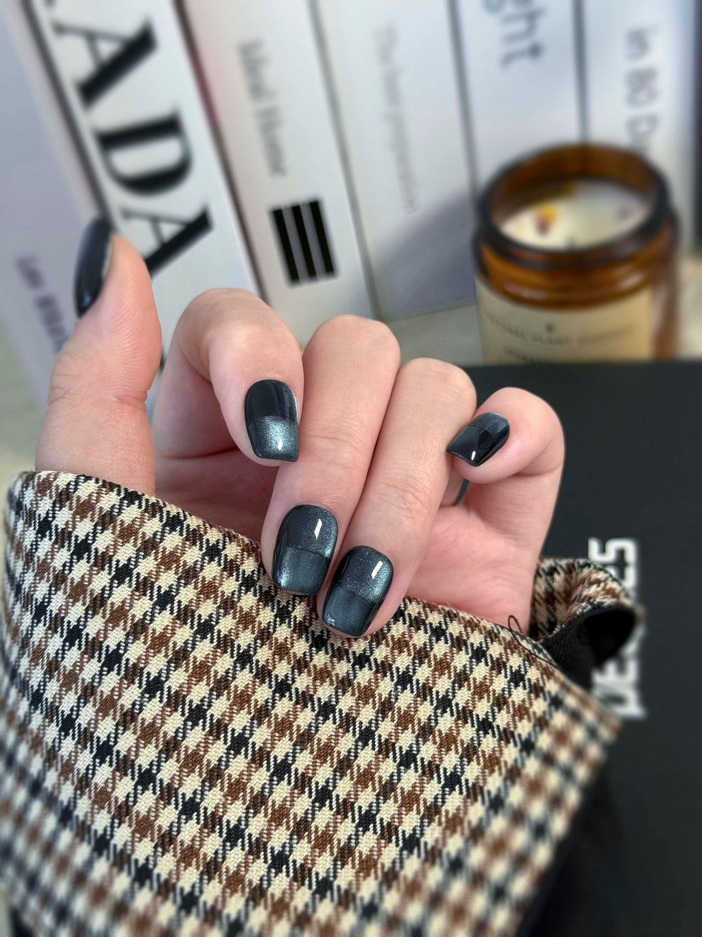 Hand with navy nail polish holding a textured fabric item, with books and a candle in the background.