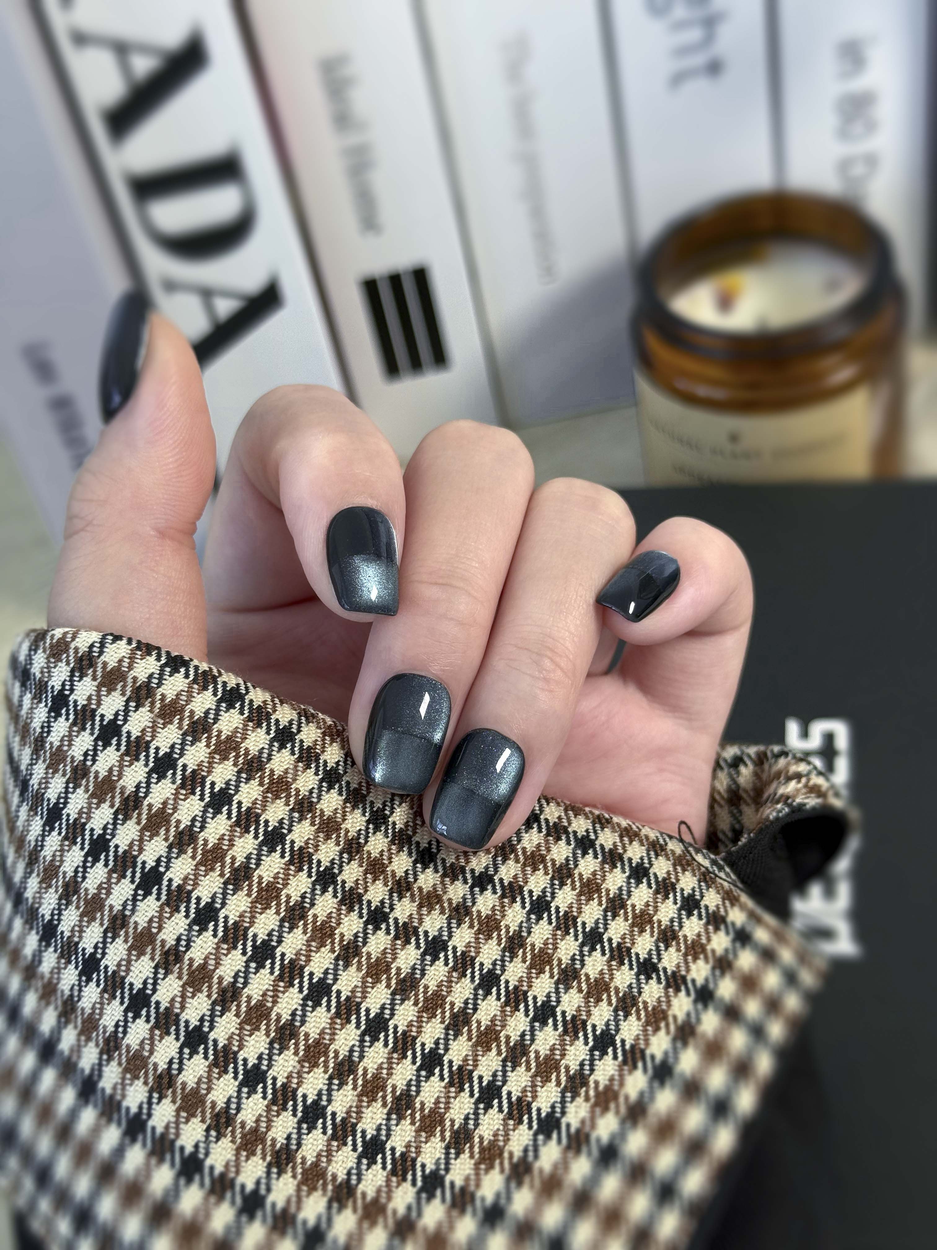 Hand with navy nail polish holding a textured fabric item, with books and a candle in the background.