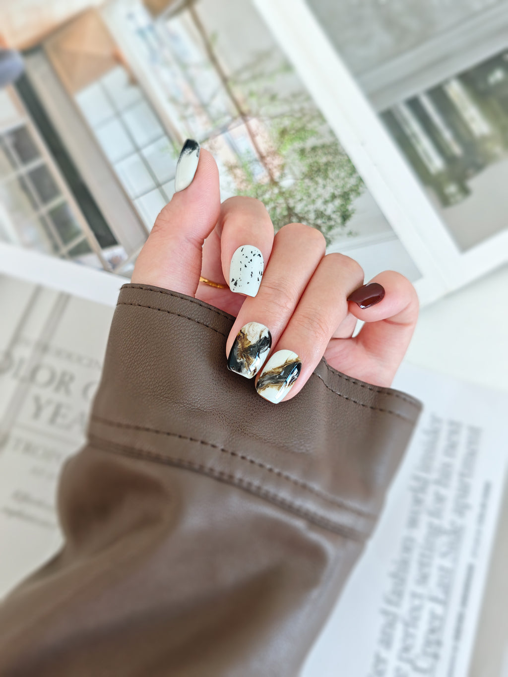 Hand with stylish inkstroke nail design holding a brown sleeve against a blurred indoor background