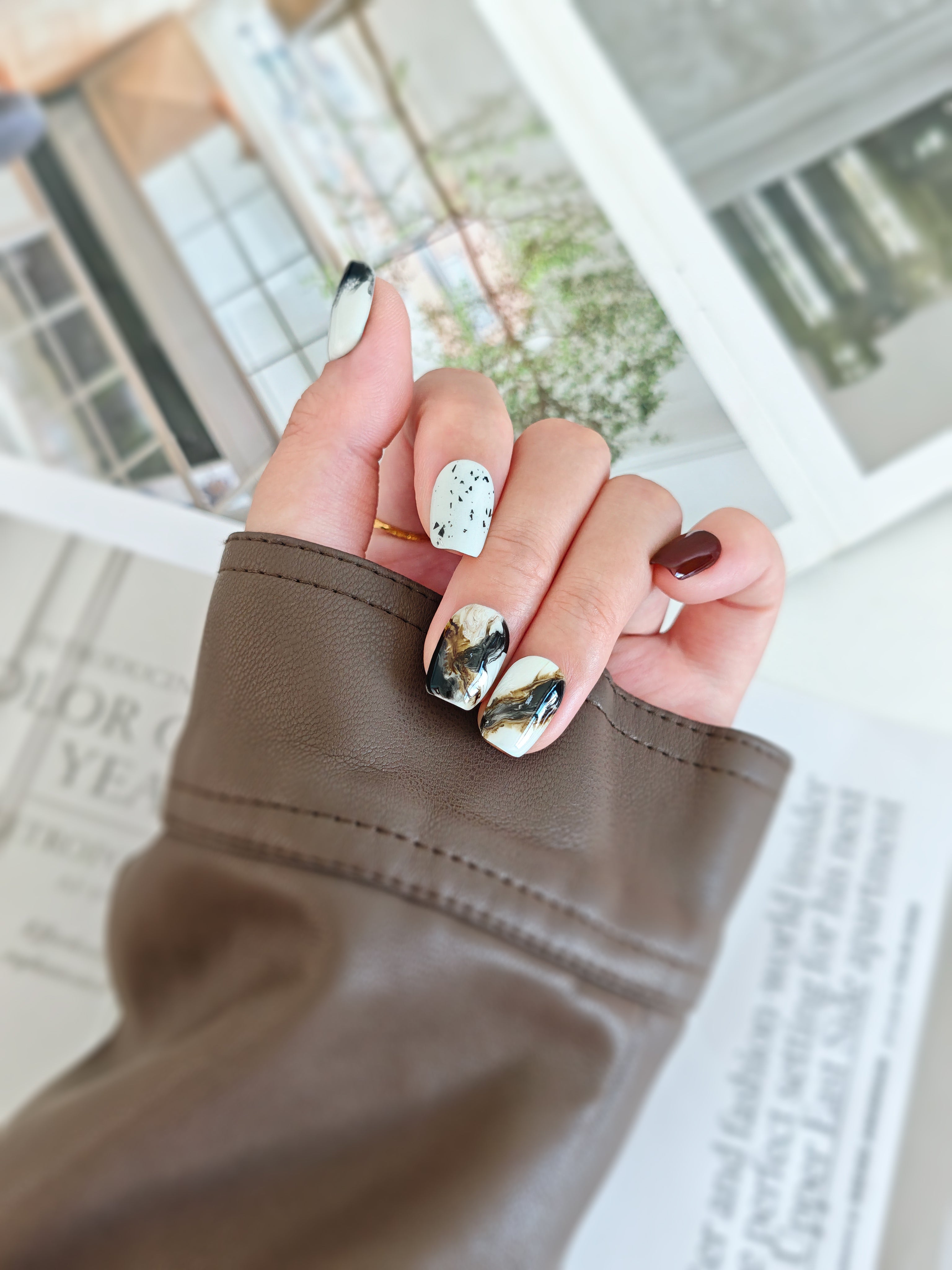 Hand with stylish inkstroke nail design holding a brown sleeve against a blurred indoor background